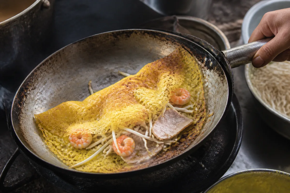 Temporary editorial illustration of a small banh xeo cooking until crisp in a hot pan