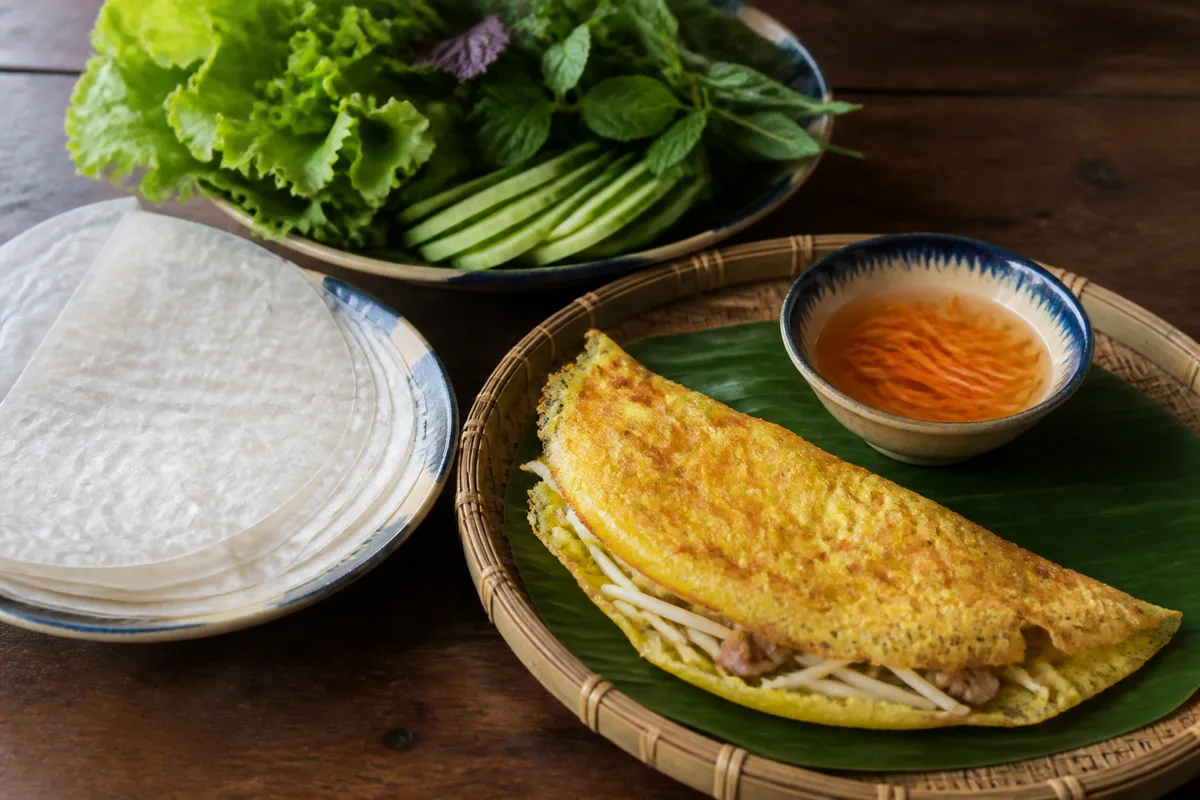 Temporary editorial illustration of banh xeo with separate rice paper, fresh herbs, cucumber and sauce