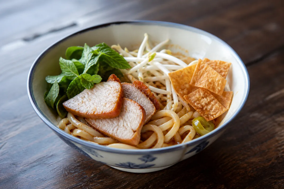 Temporary editorial illustration showing thick cao lau-style noodles, pork, herbs, sprouts and crisp rice pieces